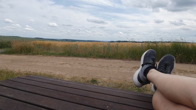 Person entspannt auf einer Bank mit Blick auf ein Feld und bewölkten Himmel.