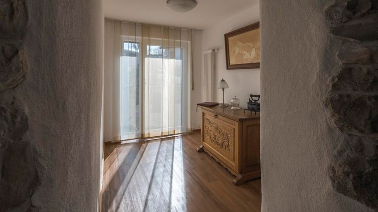 Ein heller Raum mit Holzboden und Fenster mit Vorhängen. Ein in Holz gehaltenes Möbelstück steht im Vordergrund.