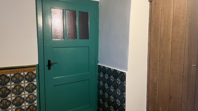 Een groene deur met ramen staat in een gang. De vloer is betegeld en aan de muur bevindt zich een decoratieve tegel.