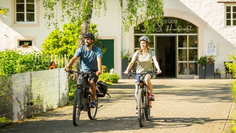 Zwei Personen fahren mit Fahrrädern auf einem gepflasterten Weg vor einem Gebäude mit der Aufschrift 'Landhofcafé'.