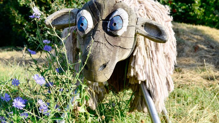 Un mouton décoratif en bois avec une expression amusante se tient dans l'herbe. Il est entouré de fleurs colorées et d'une végétation verte.