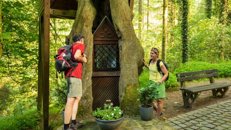 Zwei Personen betrachten eine kleine Kapelle in einem Baumstamm im Wald.