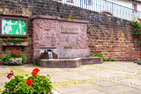 Een stenen dorpsfontein met reliëfs en stromend water, omringd door rode bloemen en een informatiebord op een stenen muur.