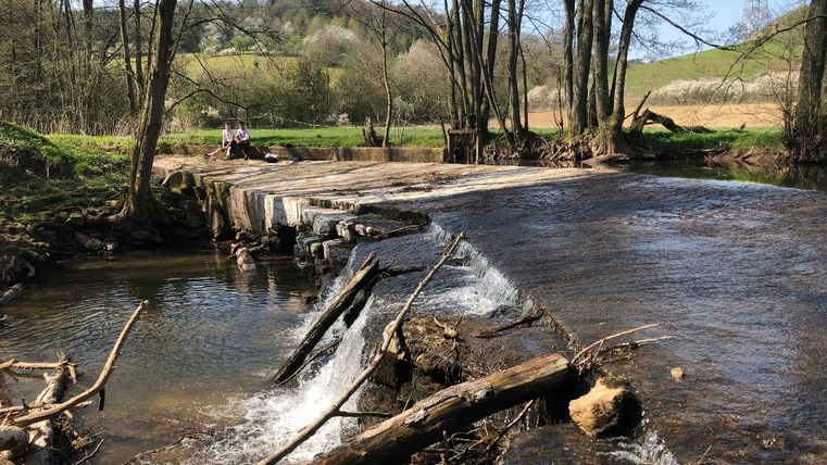 Zwei Personen sitzen auf einer kleinen Brücke über einem Fluss in einer ländlichen Landschaft.