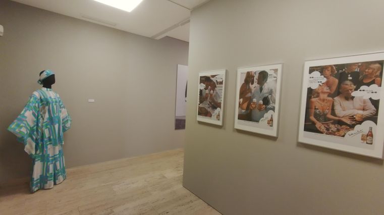 An art space with gray walls and a wooden floor. Several framed photos hang on the wall, while a figure in traditional clothing stands in the background.