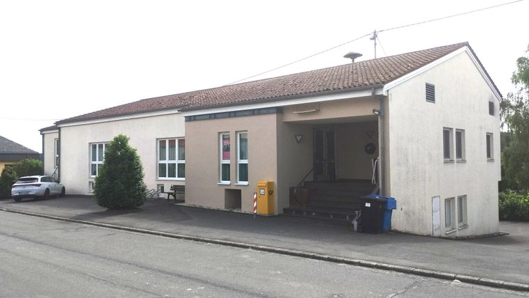 A simple building with a white facade and a red roof. In front of the building, there are some trees and parked cars.