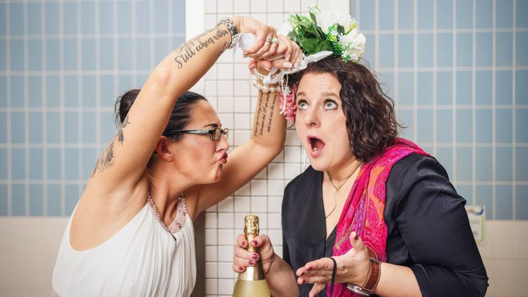 Twee vrouwen in een badkamer, de een spuit champagne en de ander kijkt verrast. De sfeer is vrolijk en feestelijk.