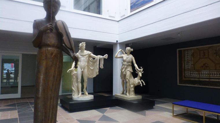 Eine große Statue eines Mannes steht neben zwei weißen Statuen, die Figuren aus der Antike darstellen. Der Raum hat eine moderne Atmosphäre mit einem klaren Design.