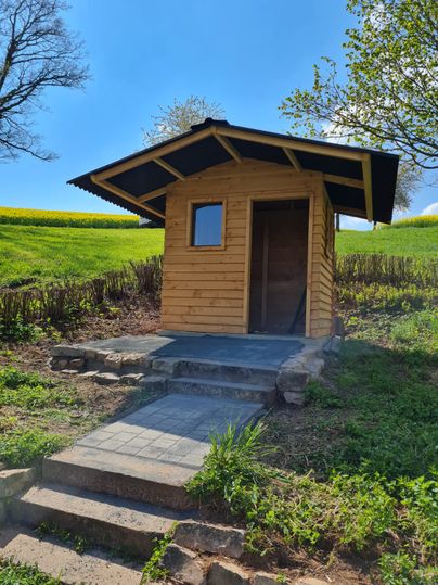 Kleines Holzhaus auf einem Hügel mit gepflastertem Weg und Treppe, umgeben von grüner Wiese und Bäumen.