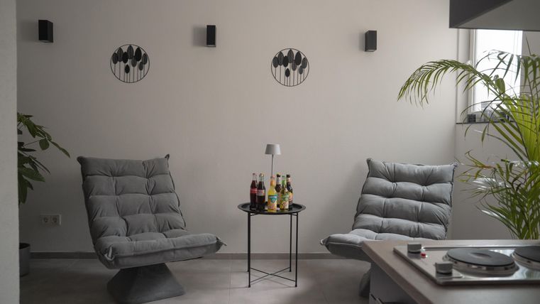 Ein gemütlicher Raum mit zwei grauen Sesseln und einem kleinen Tisch. An der Wand hängen dekorative Bilder und eine Pflanze steht in der Ecke.