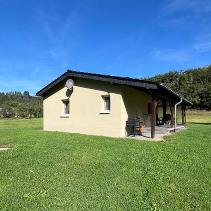 Ein einfaches Haus auf einem grünen Grundstück. Im Hintergrund sind Bäume und ein blauer Himmel zu sehen.