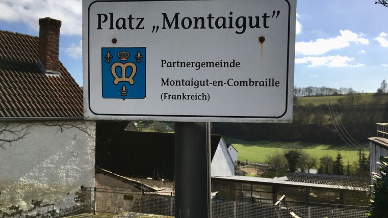 Panneau avec l'inscription 'Place Montaigut' et armoiries, paysage en arrière-plan.