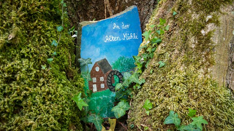 Bemalter Stein mit Mühlenmotiv und Schriftzug in Baumrinde eingebettet.