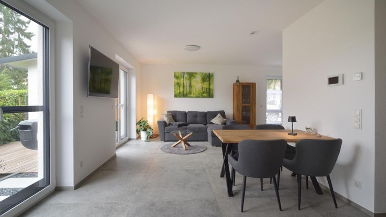 Ein modernes Wohnzimmer mit einem gemütlichen Sofa und einem Esstisch. Große Fenster sorgen für viel Tageslicht und eine entspannte Atmosphäre.