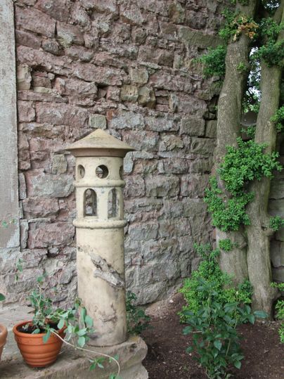 Une vieille pozzolane avec uneTour décorative se dresse contre un mur en pierre. À côté, on peut voir quelques pots de fleurs et des plantes vertes.
