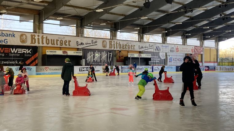 Eine Eishalle mit Kindern, die das Schlittschuhlaufen üben. Einige Kinder nutzen Schlitten zur Unterstützung.