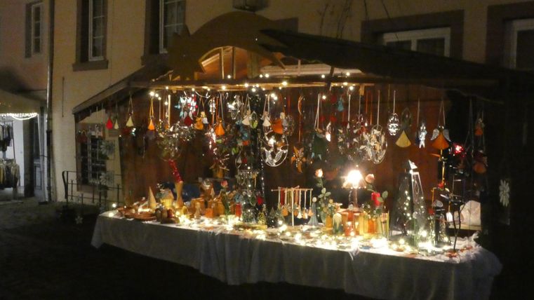 Ein festlich dekorierter Weihnachtsmarktstand mit verschiedenen handgefertigten Ornamenten und Lichtern. Die Atmosphäre ist warm und einladend.