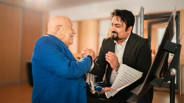 An older man in a blue suit and a younger man in a suit are smiling at each other and holding hands. In the background, blurred furniture and a sheet of music can be seen.