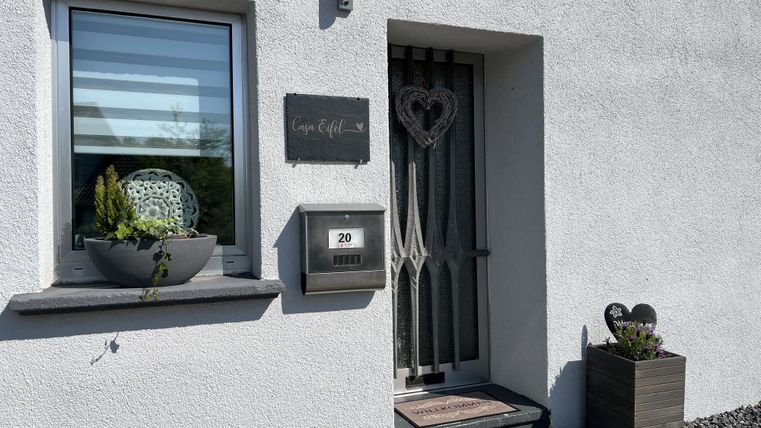 Ein Eingangsbereich mit einer weißen Wand und einer dekorativen Tür. Neben der Tür befindet sich ein Blumenkübel und ein Briefkasten.