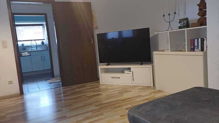 A cozy living room with a television and a modern TV stand. The room has a light wooden floor and leads to a kitchen.