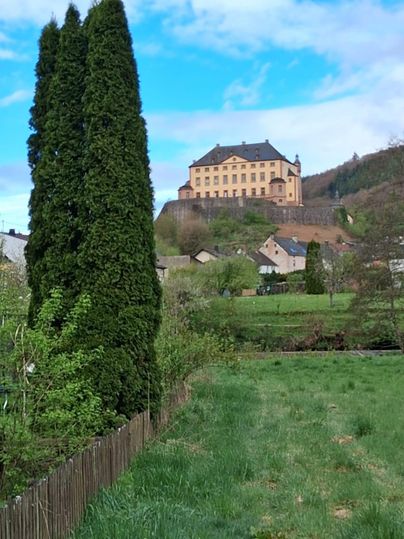 Ein historisches Schloss steht auf einem Hügel, umgeben von grünen Wiesen und Bäumen. Im Vordergrund ist ein gepflegter Garten mit einem Zaun sichtbar.