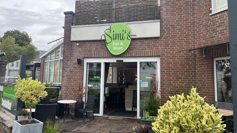 Un restaurant confortable nommé "Simi's Eat & Meet". L'entrée est entourée de plantes et de places assises.