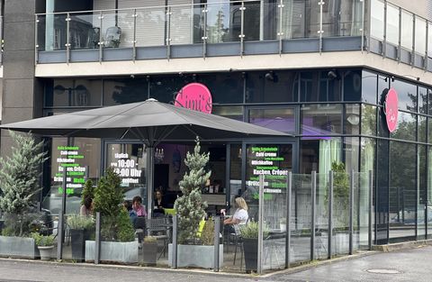 Ein modernes Café mit großen Fenstern und einer Terrasse. Es gibt Sitzplätze im Freien und dekorative Pflanzen.
