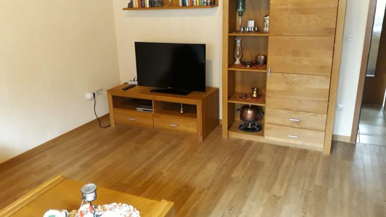 Ein gemütliches Wohnzimmer mit einem Fernseher und einem Regal aus Holz. Der Boden ist aus Holz und es gibt einen kleinen Tisch mit einer dekorativen Tischdecke.