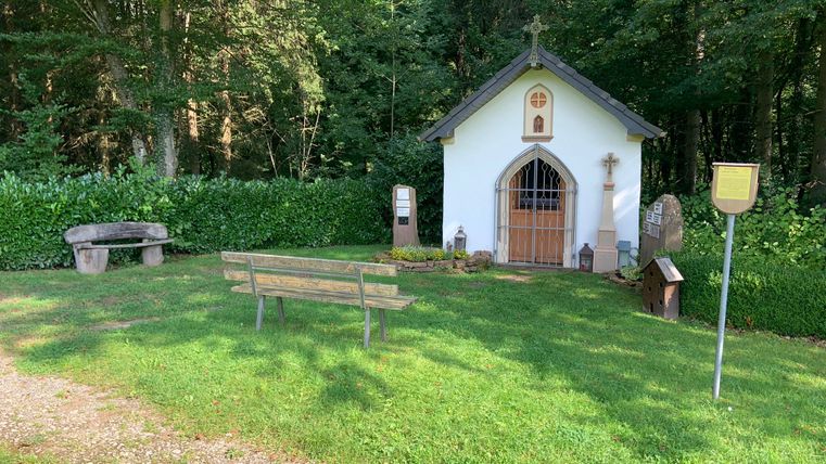 Kleine Kapelle im Wald mit Bank und Infotafel.
