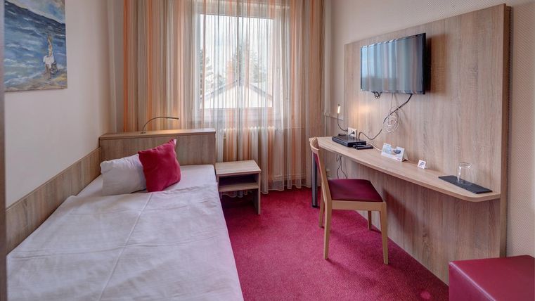 Ein einfaches Zimmer mit einem Einzelbett und einem Schreibtisch. Große Fenster lassen viel Licht herein und sorgen für eine helle Atmosphäre.