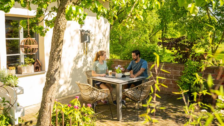 Zwei Personen sitzen an einem Tisch im Garten, umgeben von grünen Pflanzen und Bäumen.