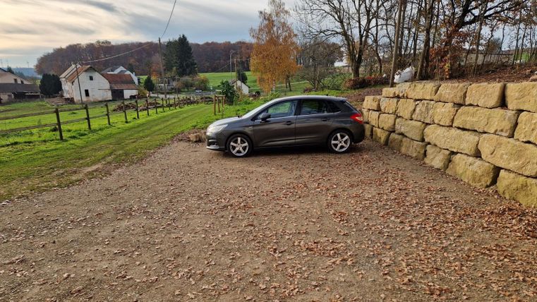 Une voiture est garée sur un parking en gravier dans un environnement rural. À l'arrière-plan, on peut voir des prairies et quelques bâtiments.