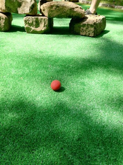 Ein roter Ball liegt auf grünem Kunstgrass neben einigen Steinen. Es ist eine ruhige Minigolf-Umgebung.