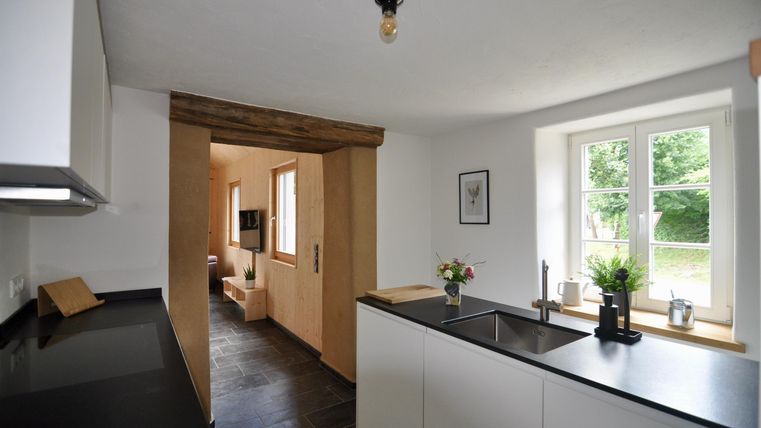 A modern kitchen with a black countertop and white cabinets. In the background, there are windows overlooking the outside and a cozy living area visible.
