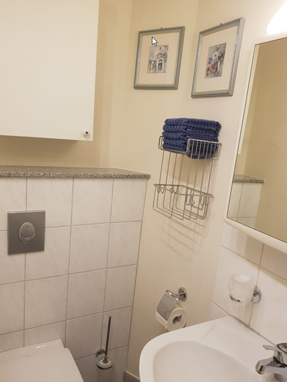 Ein kleines Badezimmer mit weißen Fliesen und einem Waschbecken. An der Wand hängen zwei Bilder und eine Regale mit Handtüchern.