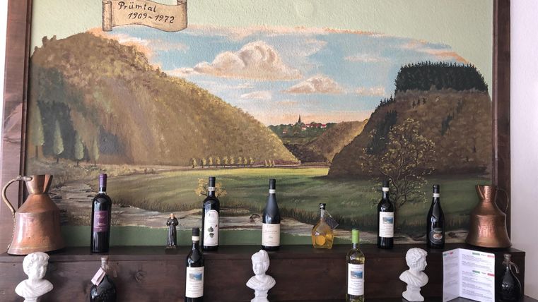 Ein Wandgemälde mit einer ländlichen Landschaft, umgeben von Hügeln und einem blauen Himmel. Darunter steht eine Holzregal mit verschiedenen Weinflaschen und dekorativen Elementen.