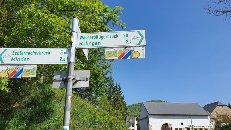 Wegweiser mit Entfernungsangaben zu Orten auf einer Radtour, umgeben von Bäumen und einem Gebäude im Hintergrund.