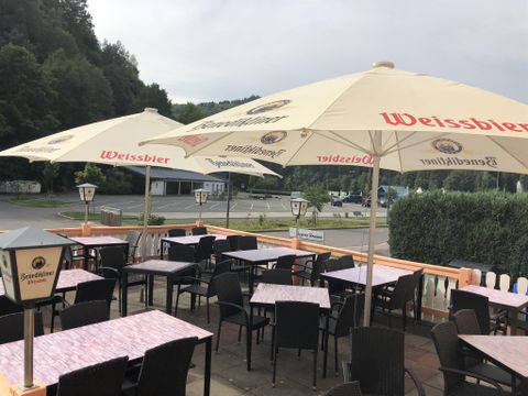 Eine Terrasse mit Tischen und Stühlen, überdacht von großen Sonnenschirmen. Im Hintergrund sind Bäume und eine ruhige Straße zu sehen.