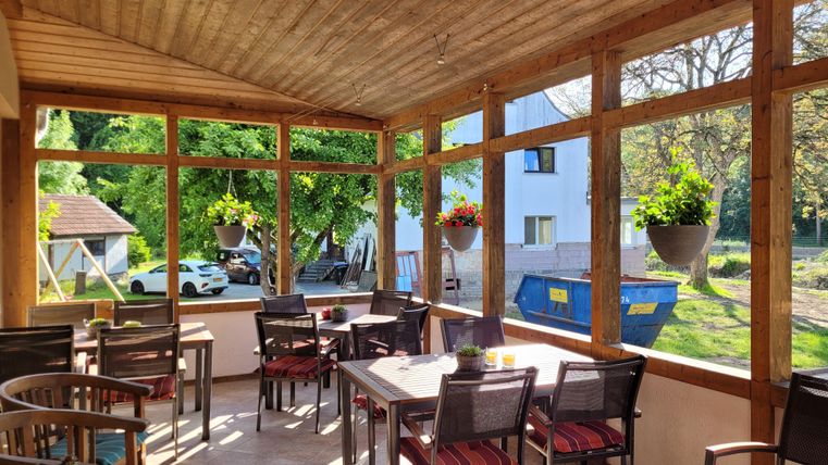 Ein schöner Wintergarten mit Holzwänden und großen Fenstern. Mehrere Tische und Stühle sind aufgestellt, umgeben von Pflanzen und viel Tageslicht.