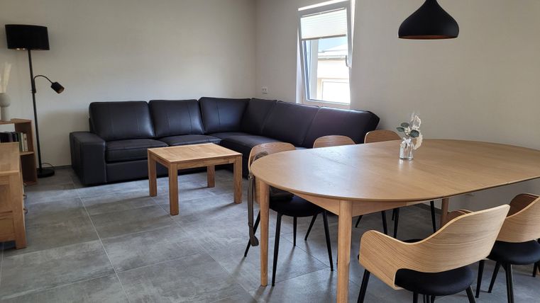 Ein modernes Wohnzimmer mit einem großen, bequemen Sofa und einem runden Esstisch. Die Einrichtung ist schlicht und einladend, mit einer Lampe und einem Fenster für natürliches Licht.