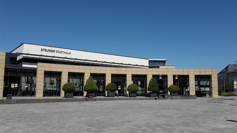 Ein modernes Gebäude mit großen Fenstern und einem klaren blauen Himmel. Vor dem Gebäude sind gleichmäßig angeordnete Bäume und Pflastersteine.