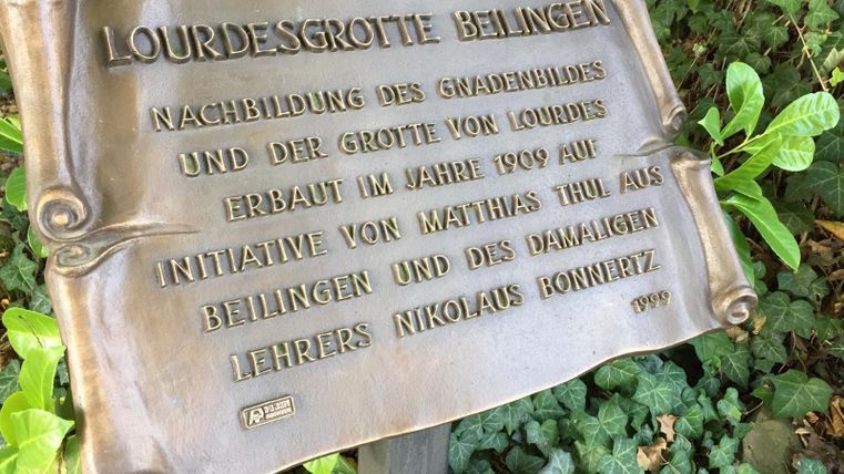 Eine bronzene Gedenktafel mit der Aufschrift „Lourdesgrotte Bellingen“. Sie informiert über die Nachbildung der Grotte von Lourdes, erbaut im Jahr 1909.