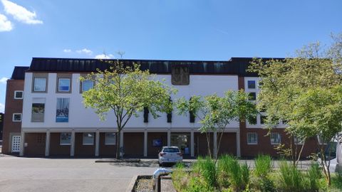 A modern building with several windows and a bright facade. Surrounding it are trees and a parking lot.