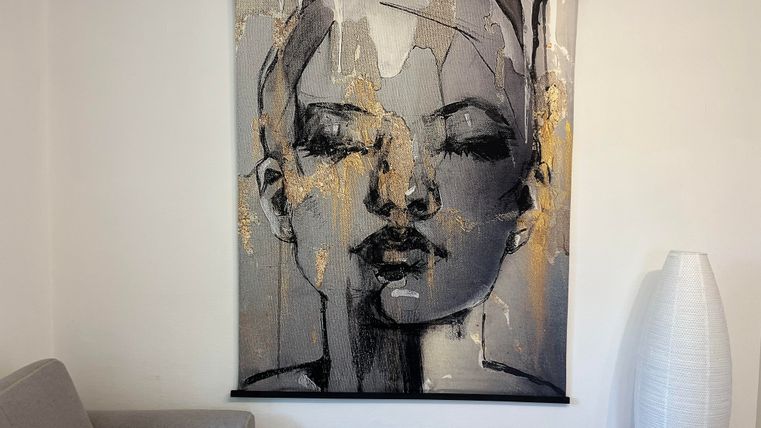 Een moderne mu mural met een abstract portret van een vrouw in zwart-wit tinten en gouden accenten. Het hangt aan een muur naast een grijze bank en een witte vloerlamp.
