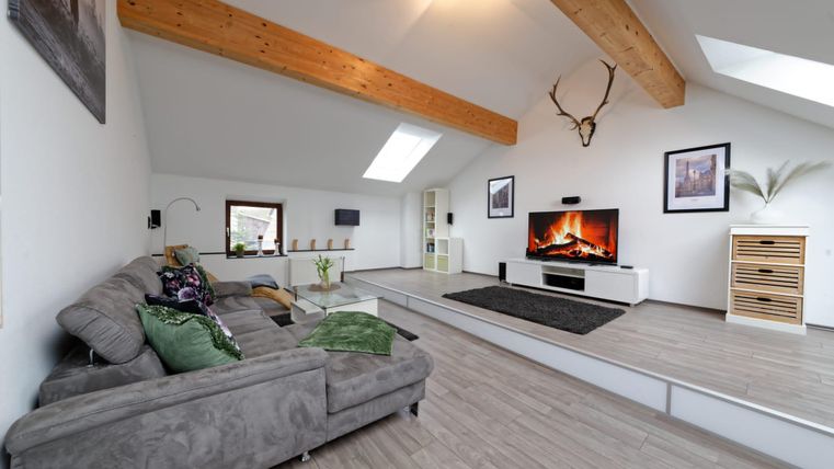 Ein modernes Wohnzimmer mit einer grauen Couch und einem großen Flachbildfernseher. Großzügige Fenster und Holzbalken verleihen dem Raum eine helle, einladende Atmosphäre.