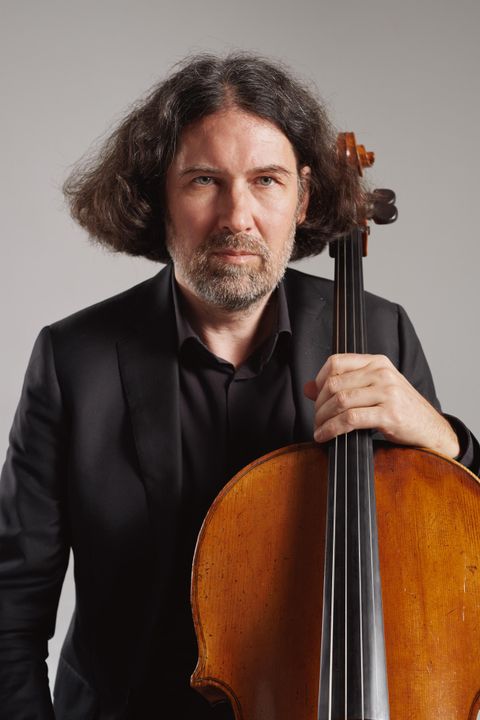 Ein Musiker mit einem Cello steht vor einem neutralen Hintergrund. Er trägt einen schwarzen Anzug und hat lockige Haare.