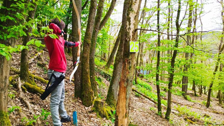 Eine Person schießt mit einem Bogen im Wald. Die Umgebung ist grün und lädt zur aktiven Erholung ein.