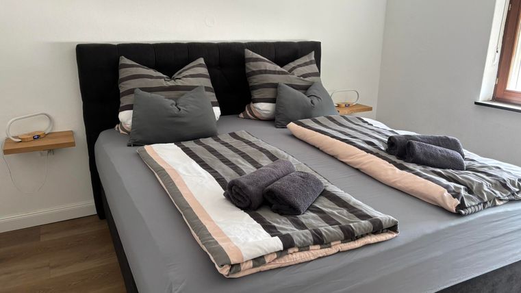 Une chambre moderne avec un grand lit double et des coussins rayés. La literie est en gris et beige, et il y a deux serviettes sur le lit.