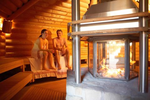 Une sauna confortable avec une lumière chaude et des panneaux en bois. Deux personnes sont assises détendues sur les bancs à côté d'une cheminée.