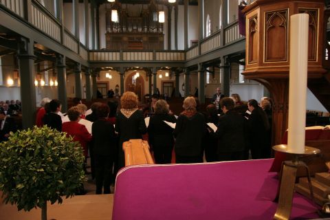 Ein Chor, der in einer Kirche singt, ist im Vordergrund zu sehen. Im Hintergrund sind die Bänke und die Kirche mit ihrer schönen Architektur zu erkennen.